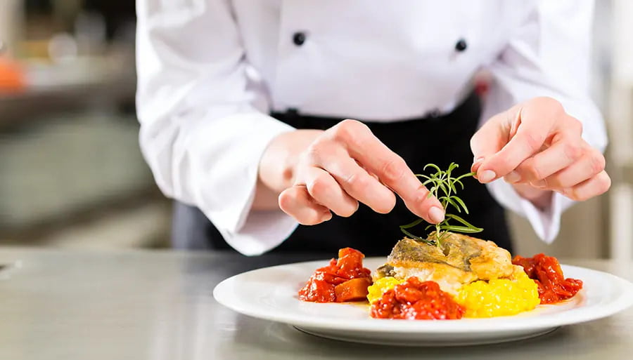 A hotel chef in a white uniform carefully garnishes a gourmet dish, showcasing the high-quality food standards of hotel businesses. The vibrant presentation reflects culinary expertise, freshness, and attention to detail, enhancing the guest dining experience.