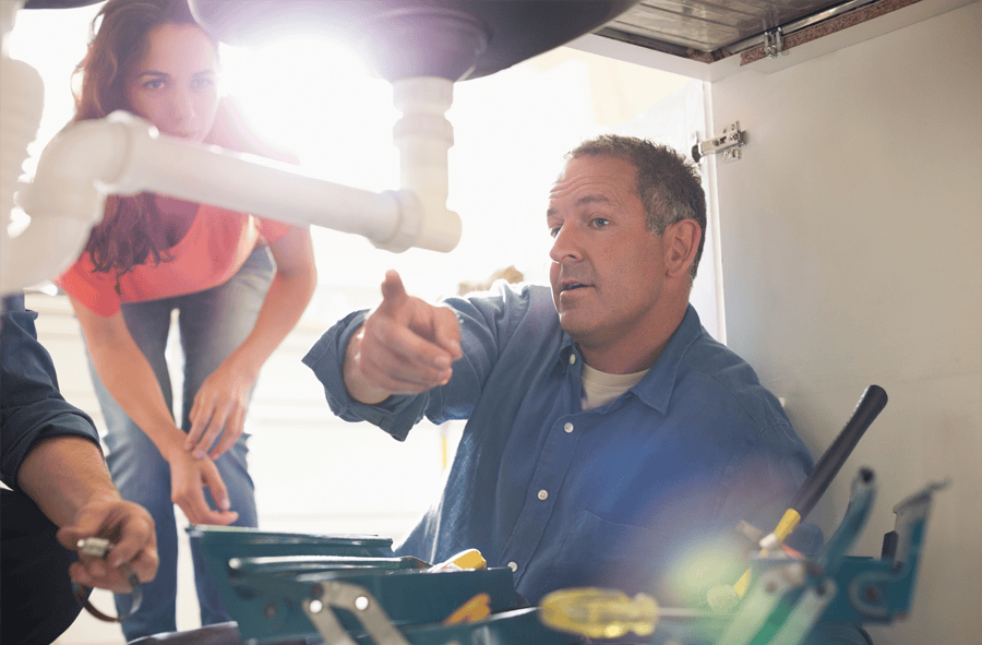 A reliable and trustworthy plumber inspects a sink pipe while explaining the issue to a concerned homeowner. With a well-stocked toolbox and a hands-on approach, the image highlights professionalism, expertise, and customer-focused service in plumbing repairs and maintenance.